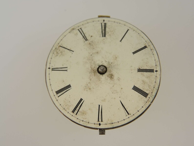 May include: A white clock face with black Roman numeral hour markers and a black minute track. The clock face is slightly dirty and has a small metal knob in the center.