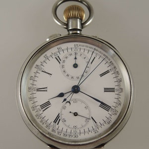 May include: A silver pocket watch with a white face and black Roman numeral hour markers. The watch has a chronograph function with a smaller dial at the bottom and a tachymeter scale around the outer edge. The watch has a gold-coloured winding stem.