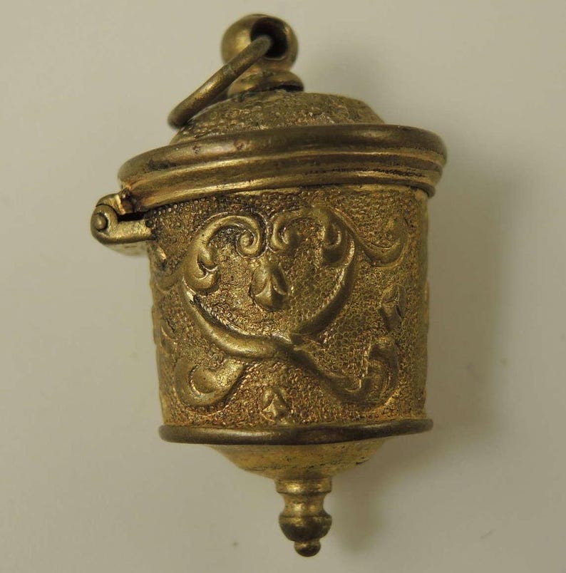 May include: A gold-toned metal thimble with an ornate design and a hinged lid. The thimble has a loop at the top for hanging.