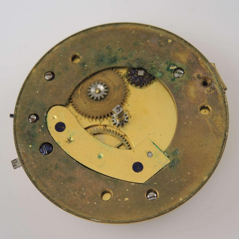 Pode incluir: Close-up de um movimento de rel&oacute;gio vintage. A placa circular de lat&atilde;o mostra engrenagens, parafusos e um balancier intrincados. O metal envelhecido tem uma p&aacute;tina com &aacute;reas de oxida&ccedil;&atilde;o verde, indicando sua idade. Vista detalhada.