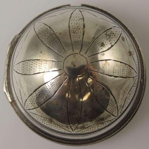 Könnte beinhalten: Ein silberner Deckel mit einem dekorativen Blumenmuster. Der Deckel ist rund und hat einen erhöhten Rand. Das Blumenmuster besteht aus acht Blütenblättern, die in einem Kreis um einen zentralen Kreis angeordnet sind.