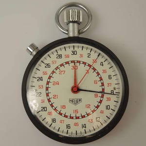 Op de afbeelding: Een zwart-witte stopwatch met een rode secondewijzer. De wijzerplaat van de stopwatch heeft het woord "HEUER" en de tekst "MADE IN SWITZERLAND".