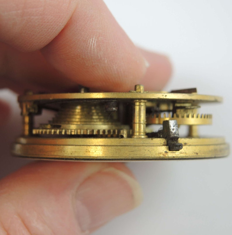 May include: Close-up side view of a brass pocket watch mechanism with gears and a winding stem. The watch is disassembled and the inner workings are visible.