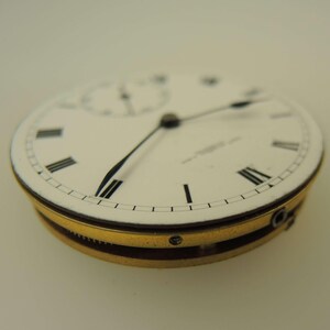 May include: Close-up of a vintage pocket watch face. The white face features black Roman numerals, black hands, and a subdial. The watch has a gold-colored bezel and a dark brown band. The watch is a classic timepiece.