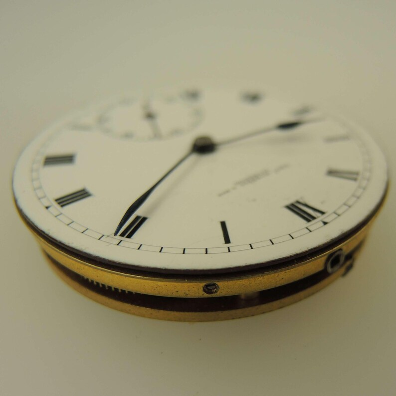 May include: Close-up of a vintage pocket watch face. The white face features black Roman numerals, black hands, and a subdial. The watch has a gold-colored bezel and a dark brown band. The watch is a classic timepiece.