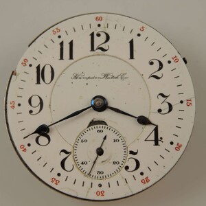 May include: A white pocket watch with black Roman numeral hour markers and black hands. The watch face has a smaller, concentric circle with black markings for seconds. The watch face also has the text "Hampden Watch Co."
