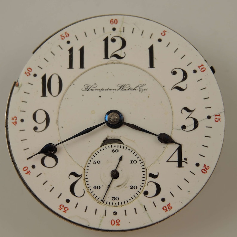 May include: A white pocket watch with black Roman numeral hour markers and black hands. The watch face has a smaller, concentric circle with black markings for seconds. The watch face also has the text "Hampden Watch Co."