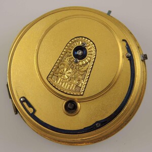 May include: A gold-colored pocket watch movement with a blue steel balance wheel and a decorative gold plate with an engraved floral design.