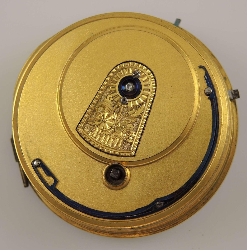 May include: A gold-colored pocket watch movement with a blue steel balance wheel and a decorative gold plate with an engraved floral design.