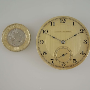 K&ouml;nnte beinhalten: Eine goldene Taschenuhr mit schwarzen Zeigern und r&ouml;mischen Ziffern. Das Zifferblatt der Uhr hat ein strukturiertes, gestreiftes Muster und die Worte "Laforge & Valentine" sind auf dem Zifferblatt aufgedruckt.