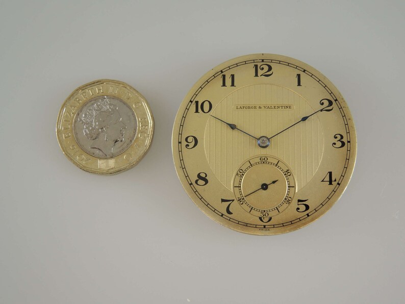 K&ouml;nnte beinhalten: Eine goldene Taschenuhr mit schwarzen Zeigern und r&ouml;mischen Ziffern. Das Zifferblatt der Uhr hat ein strukturiertes, gestreiftes Muster und die Worte "Laforge & Valentine" sind auf dem Zifferblatt aufgedruckt.