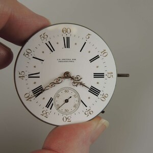 May include: A white pocket watch face with black Roman numeral hour markers and black minute markers. The watch face has a gold-colored trim and the text "C.S. SMITH & SON PHILADELPHIA." is printed on the face.