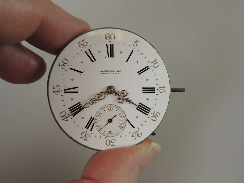 May include: A white pocket watch face with black Roman numeral hour markers and black minute markers. The watch face has a gold-colored trim and the text "C.S. SMITH & SON PHILADELPHIA." is printed on the face.