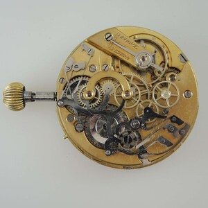 May include: A close-up view of the inner workings of a gold-toned pocket watch. The watch face is removed, revealing intricate gears and levers.