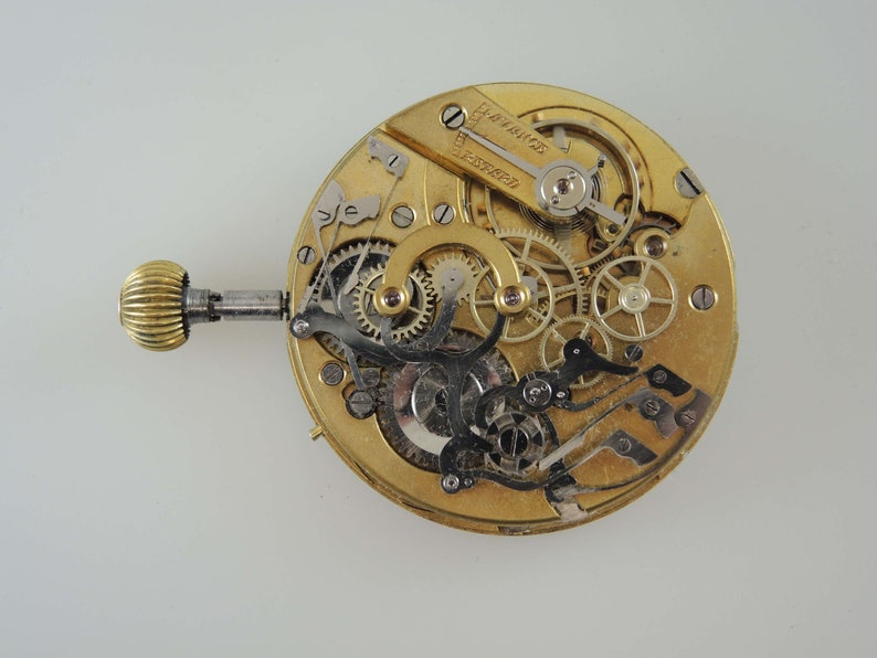 May include: A close-up view of the inner workings of a gold-toned pocket watch. The watch face is removed, revealing intricate gears and levers.