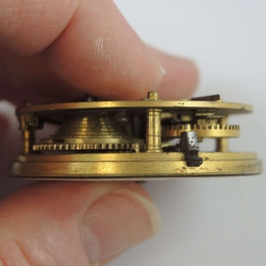 May include: Close-up side view of a brass pocket watch mechanism with intricate gears and a black metal lever.