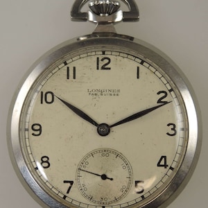 Puede incluir: Un reloj de bolsillo plateado con una esfera blanca y agujas negras. El reloj tiene un pequeño dial de segundero en la parte inferior de la esfera. La esfera del reloj tiene marcadores de hora romanos negros y marcadores de minutos arábigos negros. La esfera del reloj también tiene el texto "LONGINES FAB. SUISSE".