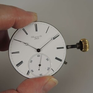 May include: A white pocket watch face with black Roman numeral hour markers and black hands. The watch face has the text "Dent & Co. London" printed on it. The watch has a gold winding stem.