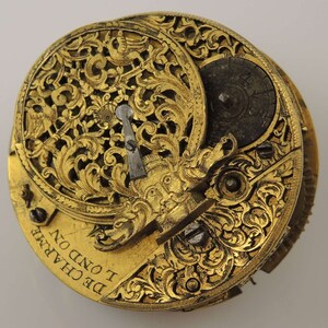 May include: A gold-toned pocket watch with intricate floral engravings and a small, round dial. The watch face is inscribed with the text "DE CHARME I OND ON".