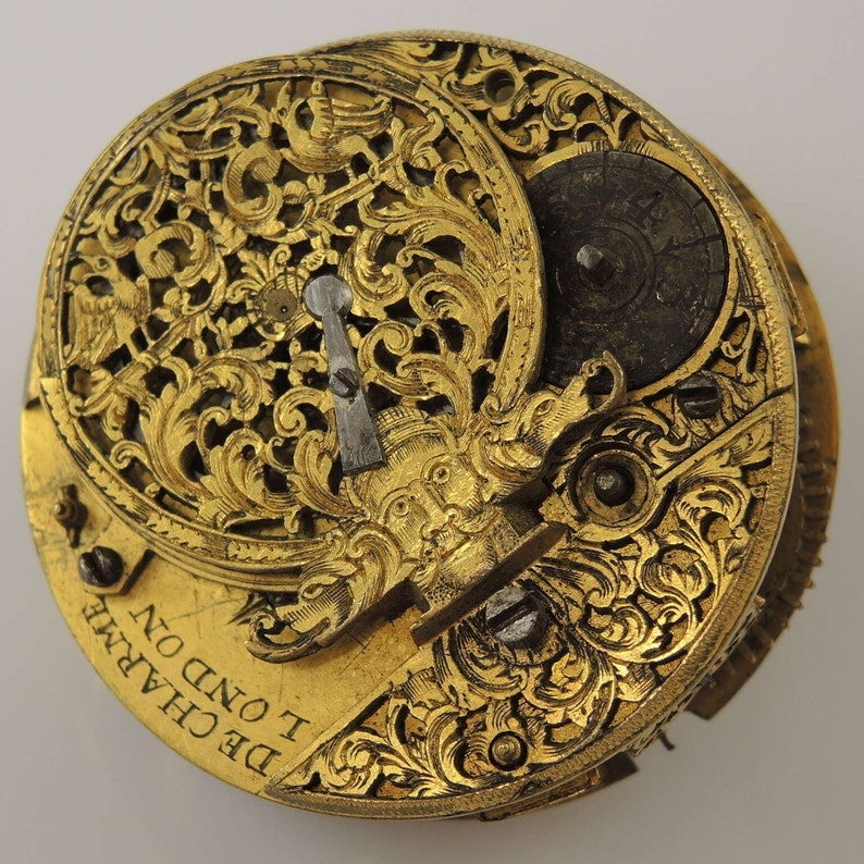 May include: A gold-toned pocket watch with intricate floral engravings and a small, round dial. The watch face is inscribed with the text "DE CHARME I OND ON".