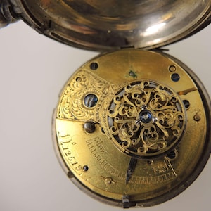 May include: Open pocket watch with a silver case and a gold-toned movement. The movement features intricate filigree and engraved details, including the number "12519". The watch is partially open, revealing the internal mechanisms.