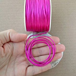 Könnte beinhalten: Eine Spule mit leuchtend pinkfarbenem gewachstem Garn, wobei eine Länge des Garns um eine weiße Pappspule gewickelt ist. Das Garn ist auch in einer separaten Spule gezeigt, die um eine Hand gewickelt ist.