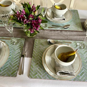 May include: A table setting with four place settings, each with a white bowl, a plate, a napkin, and a fork. The bowls contain pears. The table is covered with a light brown tablecloth and green placemats with a herringbone pattern. There are also two champagne flutes and a vase of purple flowers.