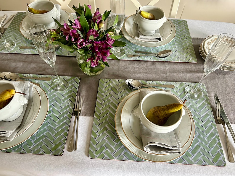 May include: A table setting with four place settings, each with a white bowl, a plate, a napkin, and a fork. The bowls contain pears. The table is covered with a light brown tablecloth and green placemats with a herringbone pattern. There are also two champagne flutes and a vase of purple flowers.