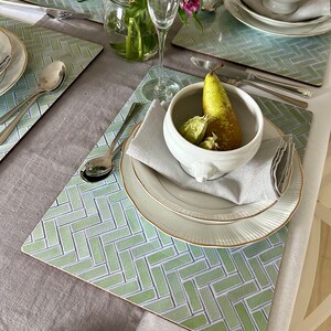 May include: A green and white placemat with a herringbone pattern. A white bowl with a pear and green fruit inside is on top of the placemat. A white plate with a gold rim is under the bowl. A white napkin is folded next to the bowl.