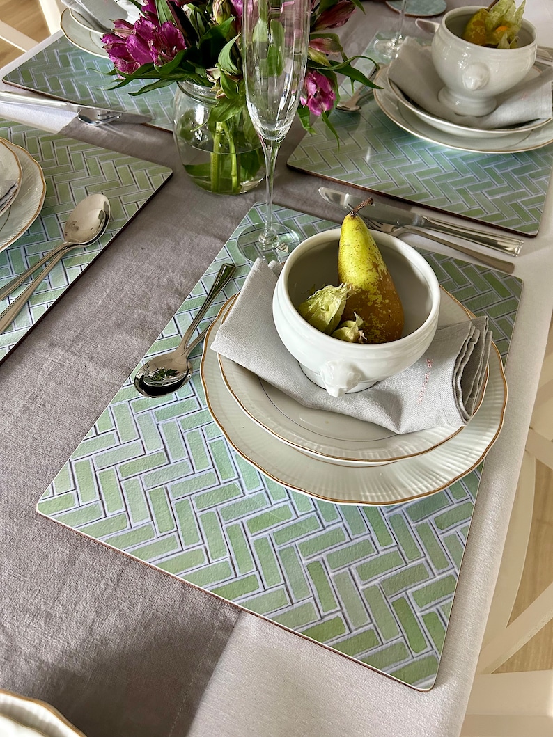 May include: A green and white placemat with a herringbone pattern. A white bowl with a pear and green fruit inside is on top of the placemat. A white plate with a gold rim is under the bowl. A white napkin is folded next to the bowl.