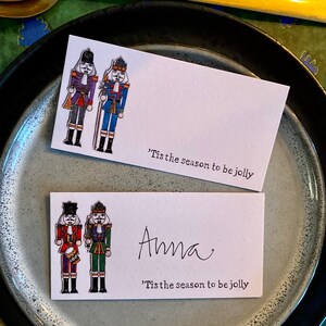 Op de afbeelding: Twee kerstkaartjes met een afbeelding van een notenkraker. De kaartjes hebben een rood en groen notenkraker en een blauw en gouden notenkraker. De tekst op de kaartjes luidt 'Tis the season to be jolly'. Op één kaartje staat de naam 'Anna'.