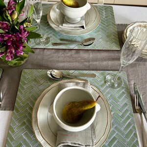 May include: Two place settings with green and white patterned placemats, white soup bowls with a pear inside, and white plates with gold trim.  A gray linen napkin is folded on each plate.