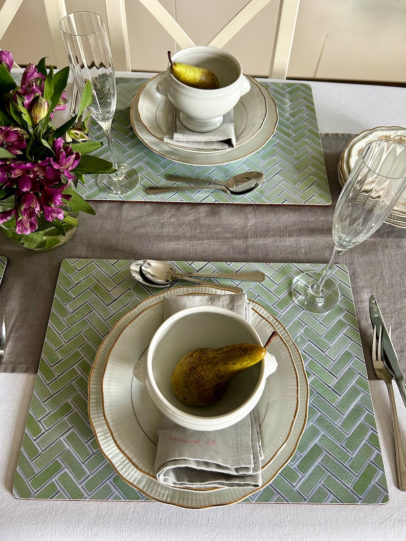 May include: Two place settings with green and white patterned placemats, white soup bowls with a pear inside, and white plates with gold trim.  A gray linen napkin is folded on each plate.