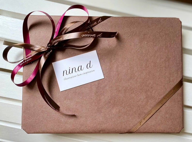 May include: A brown paper package tied with a burgundy and brown ribbon. The package is labeled with a white card that reads "nina d illustration + home inspiration".
