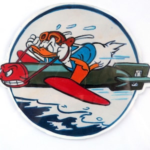 Può includere: Un adesivo circolare con un'anatra dei cartoni animati con casco e occhiali da pilota, in sella a un oggetto rosso a forma di aereo attaccato a una bomba verde. L'anatra è in volo, con un personaggio dalla faccia rossa davanti.