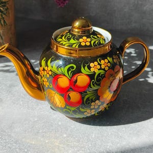 Może przedstawiać: Czarny ceramiczny czajniczek z ręcznie malowanym motywem kwiatowym w kolorach czerwonym, pomarańczowym i żółtym z zielonymi liśćmi. Czajniczek posiada błyszczącą, złotą wylewkę, uchwyt i pokrywkę oraz pasującą złotą krawędź wokół otworu.
