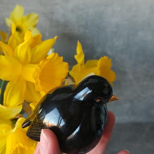 Puede incluir: Una figura de pájaro negro brillante con un pequeño pico naranja. El pájaro se sostiene en una mano, con brillantes narcisos amarillos en el fondo. La figura tiene una superficie lisa y reflectante.