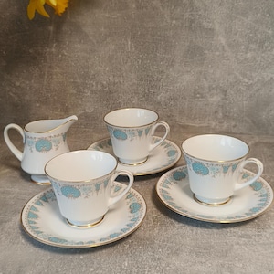 Könnte beinhalten: Ein Vintage-Teeservice mit weißen Tassen, Untertassen und einem Sahnekännchen, verziert mit einem blauen Blumenmuster und goldenen Rändern. Das Set umfasst drei Tassen, drei Untertassen und ein Sahnekännchen. Gelbe Narzissen sind im Hintergrund.