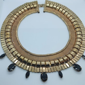 May include: A gold-coloured statement necklace with a layered design. The necklace features a mesh base, a middle layer of rectangular links, and an outer layer of larger, rounded links. Black oval-shaped gemstones dangle from the bottom.