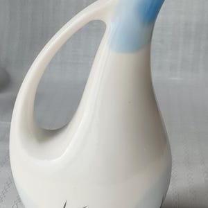 May include: White ceramic pitcher with a large handle and a blue-painted rim. The pitcher has a teardrop shape with a black arrow design on the front. A decorative piece for serving beverages.