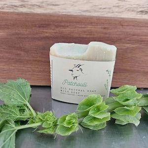 Puede incluir: Una pastilla de jabón de leche de cabra hecho a mano con una etiqueta blanca que dice "Patchouli All Natural Goat Milk Soap Hand Crafted | Made With Oils". El jabón está sobre una superficie de madera con hojas verdes.