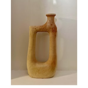 Peut inclure: Un vase en céramique beige de forme rectangulaire unique, avec une grande ouverture centrale. Le vase présente un col cylindrique et une surface légèrement texturée, avec des accents brun foncé.