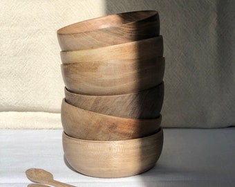May include: A stack of six wooden bowls in various sizes, all made of the same light brown wood. The bowls are stacked on a white surface, and there are three wooden spoons in front of them.