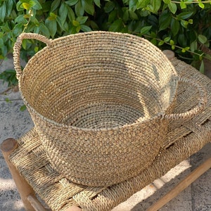 Może przedstawiać: Duży, okrągły, naturalnie pleciony kosz z dwoma uchwytami. Kosz wykonany jest z jasnobrązowego materiału i ma naturalny, rustykalny wygląd. Kosz stoi na plecionym podłożu.