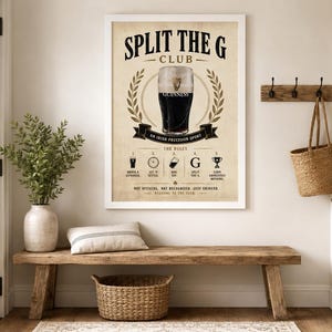 May include: A framed poster with the text "SPLIT THE G CLUB" and an illustration of a pint of Guinness. The poster is displayed above a wooden bench with a woven basket and a decorative pillow. A plant in a vase is also present.