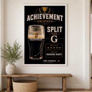Puede incluir: Un p&oacute;ster negro enmarcado con el texto "ACHIEVEMENT UNLOCKED SPLIT THE G" y una pinta de Guinness. El p&oacute;ster incluye un gr&aacute;fico de trofeo y el logotipo de Guinness. La dificultad se califica con cinco estrellas, y la recompensa es "BRAGGING RIGHTS."