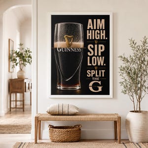 May include: A framed Guinness advertisement features a pint of stout with the brand's logo. The black background contrasts with the white frame and text: "AIM HIGH. SIP LOW. SPLIT THE G." The image is displayed above a bench with a woven basket and pillow.