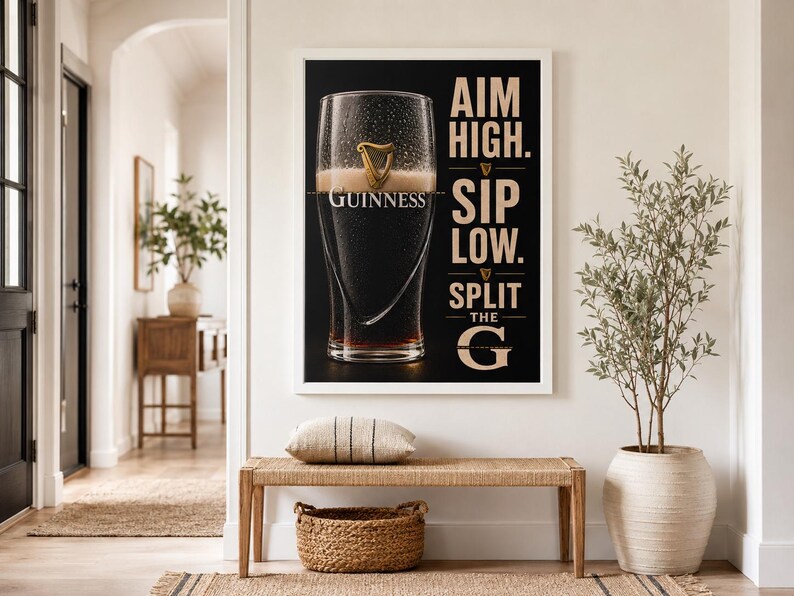 May include: A framed Guinness advertisement features a pint of stout with the brand's logo. The black background contrasts with the white frame and text: "AIM HIGH. SIP LOW. SPLIT THE G." The image is displayed above a bench with a woven basket and pillow.