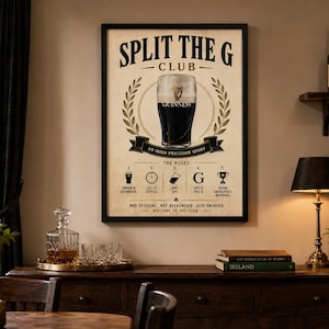 May include: A framed poster with the text "SPLIT THE G CLUB" and a pint of Guinness. The poster includes the words "AN IRISH PRECISION SPORT" and "THE RULES". The poster is displayed above a wooden cabinet with a lamp, books, and a small plant.