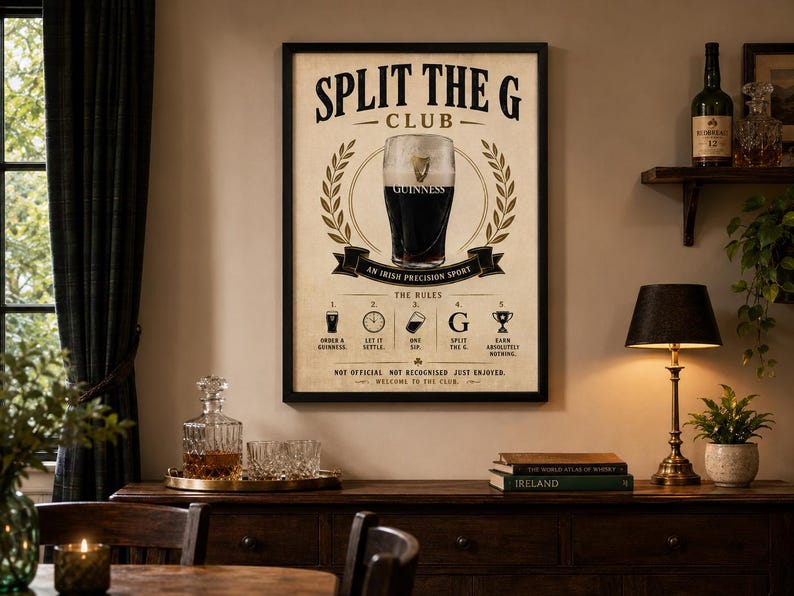 May include: A framed poster with the text "SPLIT THE G CLUB" and a pint of Guinness. The poster includes the words "AN IRISH PRECISION SPORT" and "THE RULES". The poster is displayed above a wooden cabinet with a lamp, books, and a small plant.